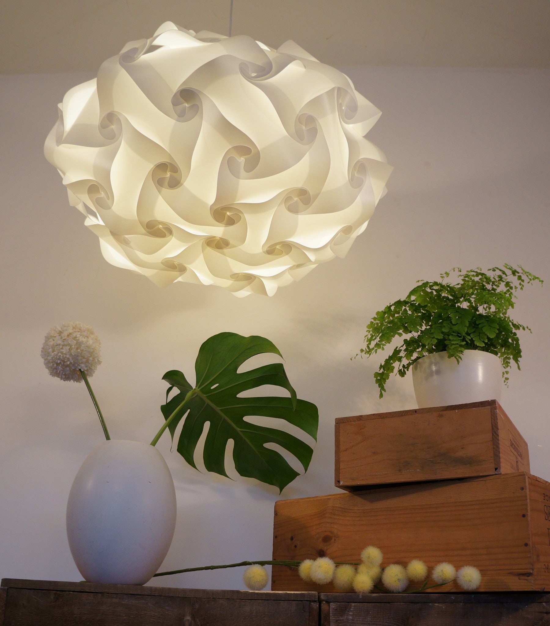 Swirling white lamp shade, vibrant green tropical leaves, decorative wooden crates, and a vase with white flowers on a rustic display shelf.