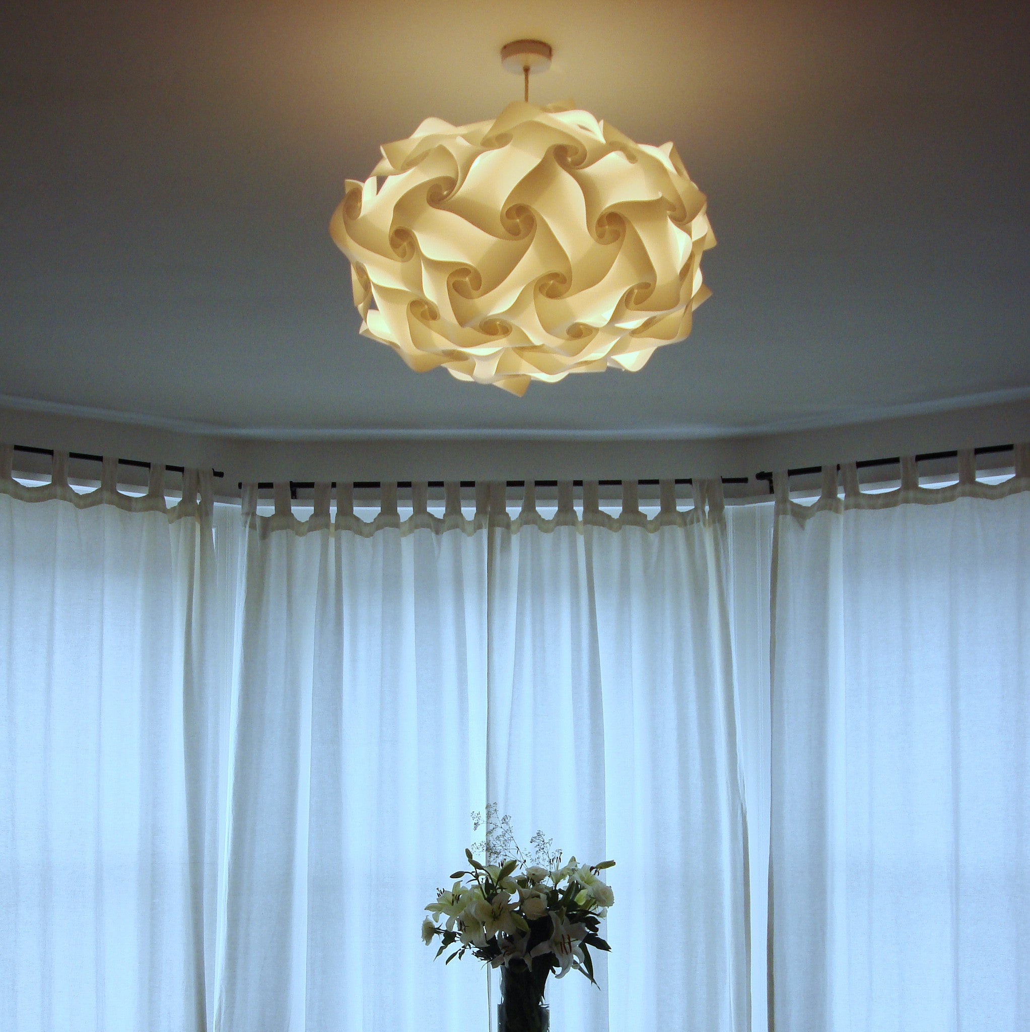 Elegant modern lampshade in an intricate, woven design made of cream-colored material, suspended from a ceiling fixture in a bright, open room with flowing curtains.