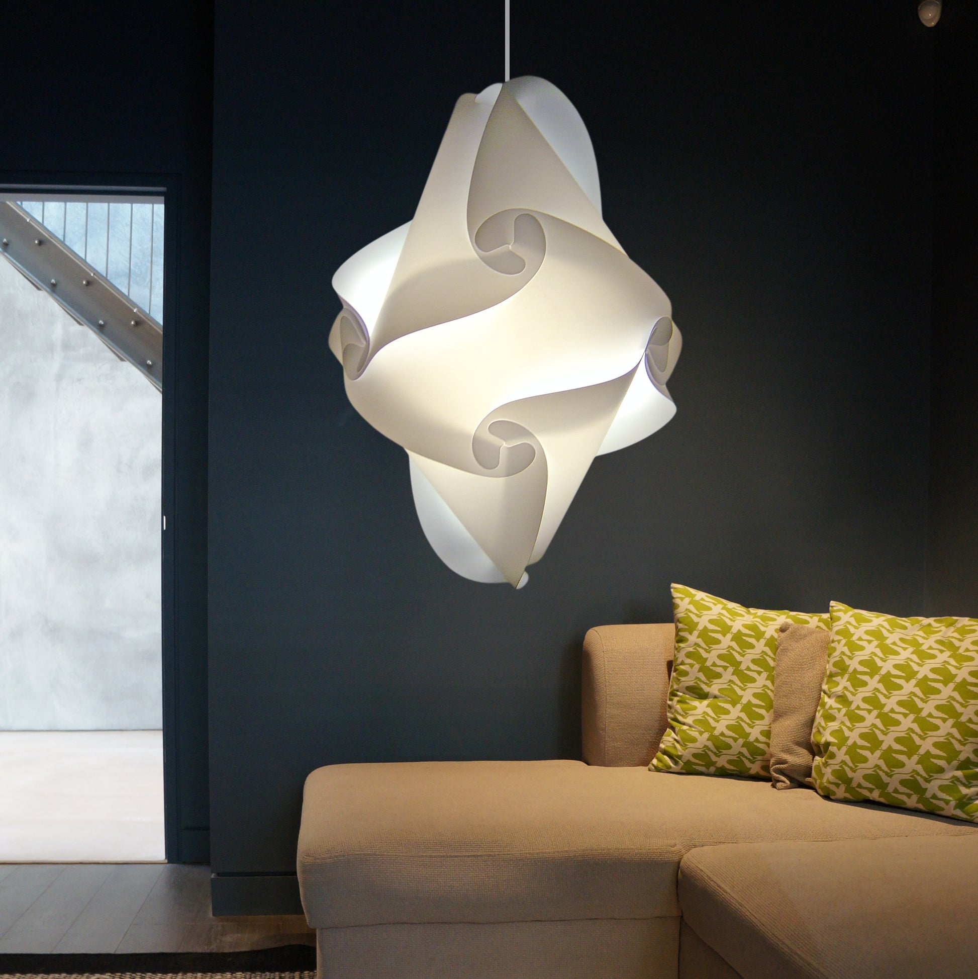 Modern white swirled lampshade suspended above a beige couch with colorful patterned cushions in a dimly lit room with a dark wall.