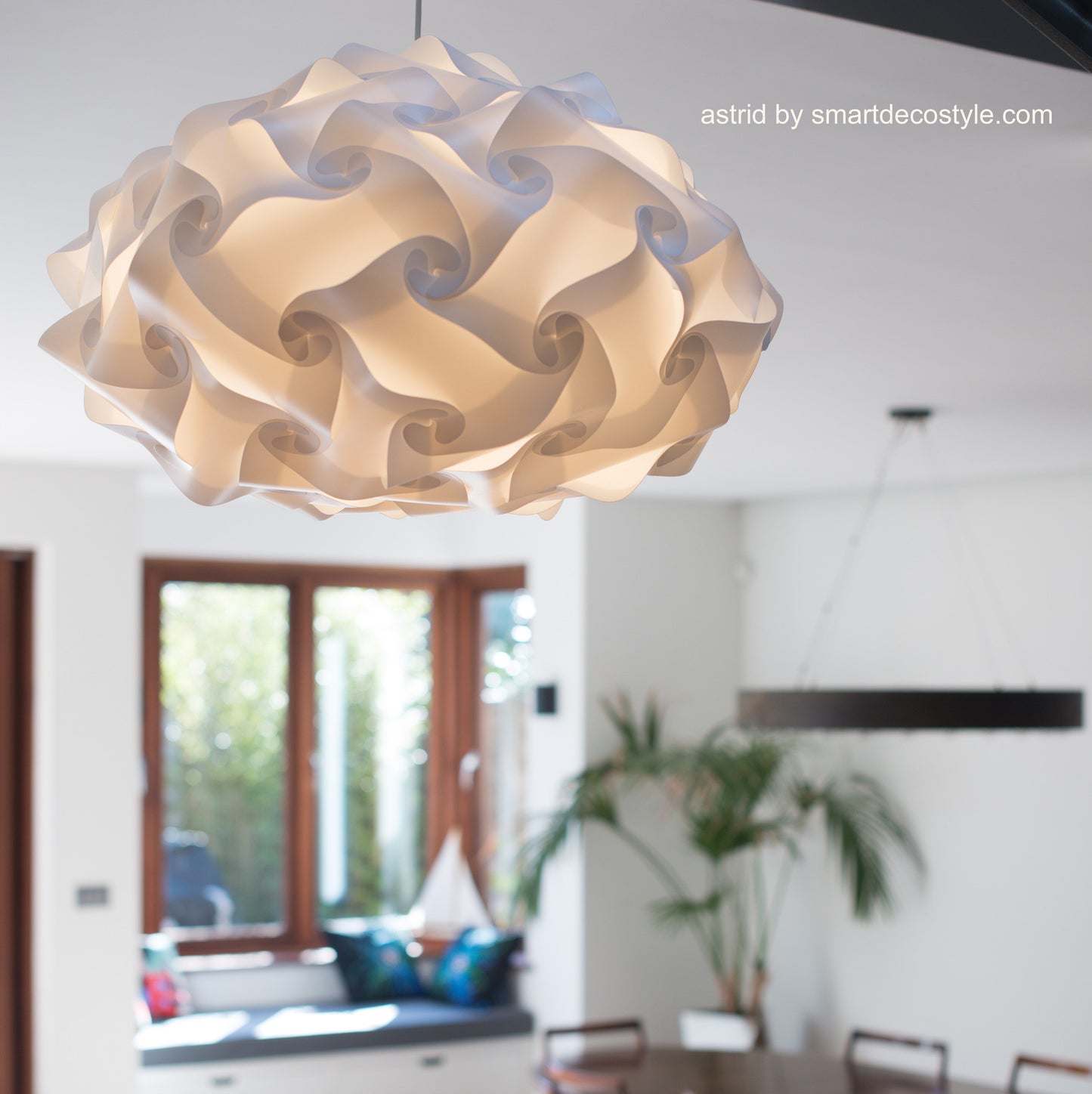 Elegant white lampshade with intricate swirls, featured in a modern interior setting with plants and windows.