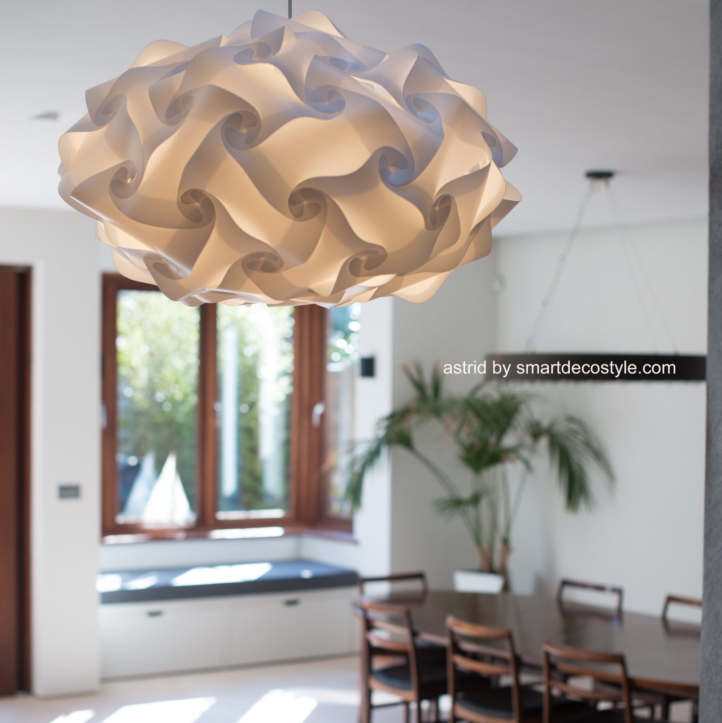 Decorative white lampshade with intricate swirl pattern hanging in a modern interior with wooden furniture and greenery.