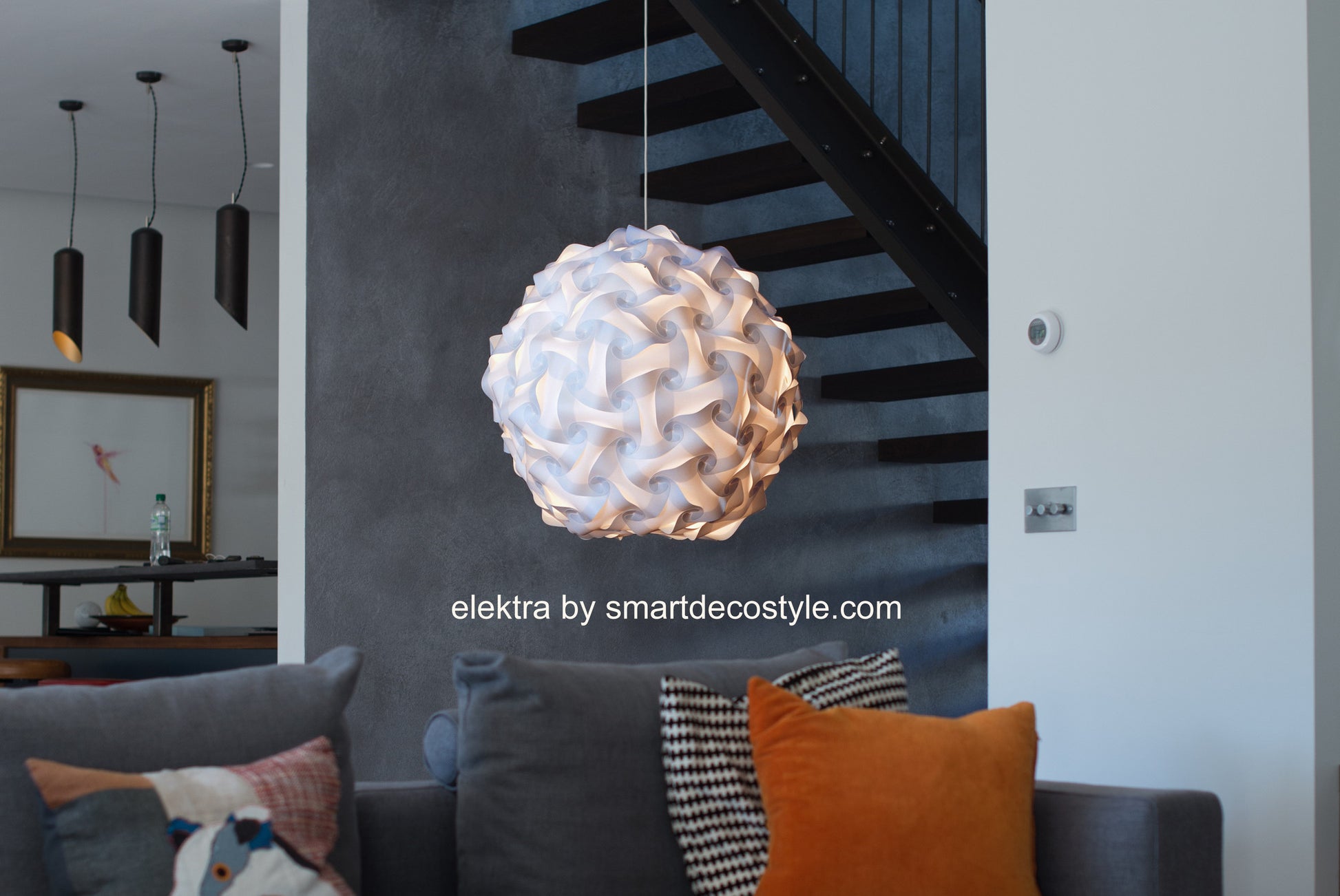 Elegant modern ceiling light shade in white, hanging above a cozy living room with dark accents and patterned throw pillows.