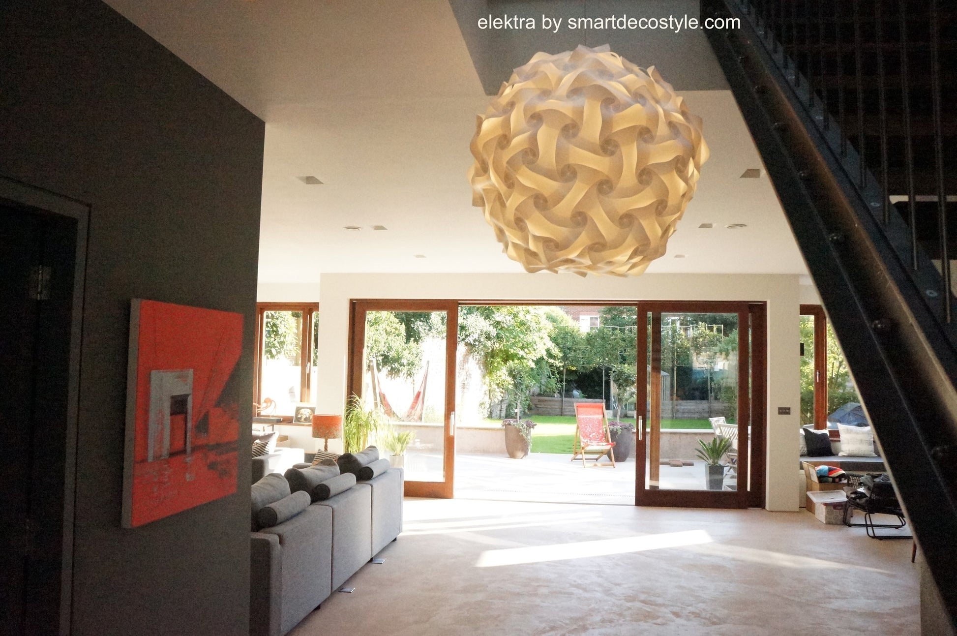 Stylish modern pendant light fixture with intricate white lampshade design suspended from ceiling in bright, open living room with large windows overlooking outdoor garden area.
