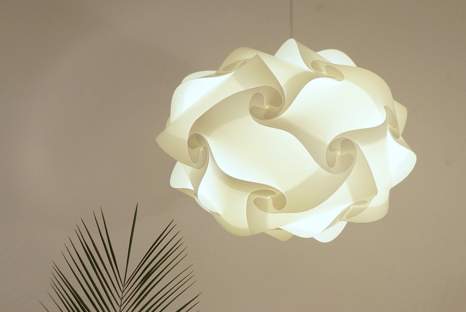 Elegant white lampshade with intricate folded design, suspended from the ceiling against a neutral background with a palm leaf accent.
