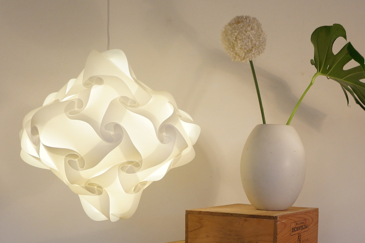 Elegant white pleated lampshade suspended from ceiling, showcasing modern design. Minimal white vase with green plant accent on wooden side table, creating a calming, contemporary home decor scene.