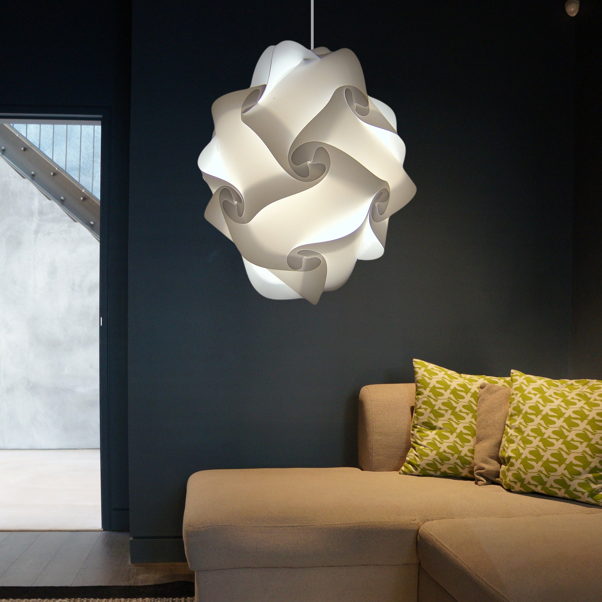 Contemporary white sculptural ceiling lampshade suspended above plush beige sofa with patterned decorative throw pillows in dark modern interior.