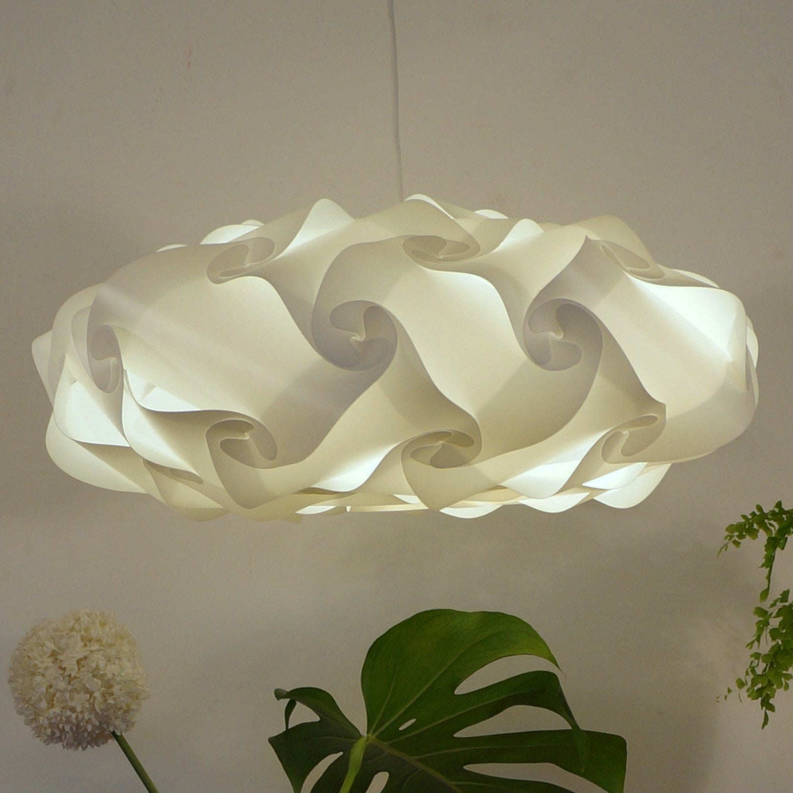 Elegant white sculptural pendant lamp with intricate wavy design, suspended in a minimalist interior setting with greenery accents.