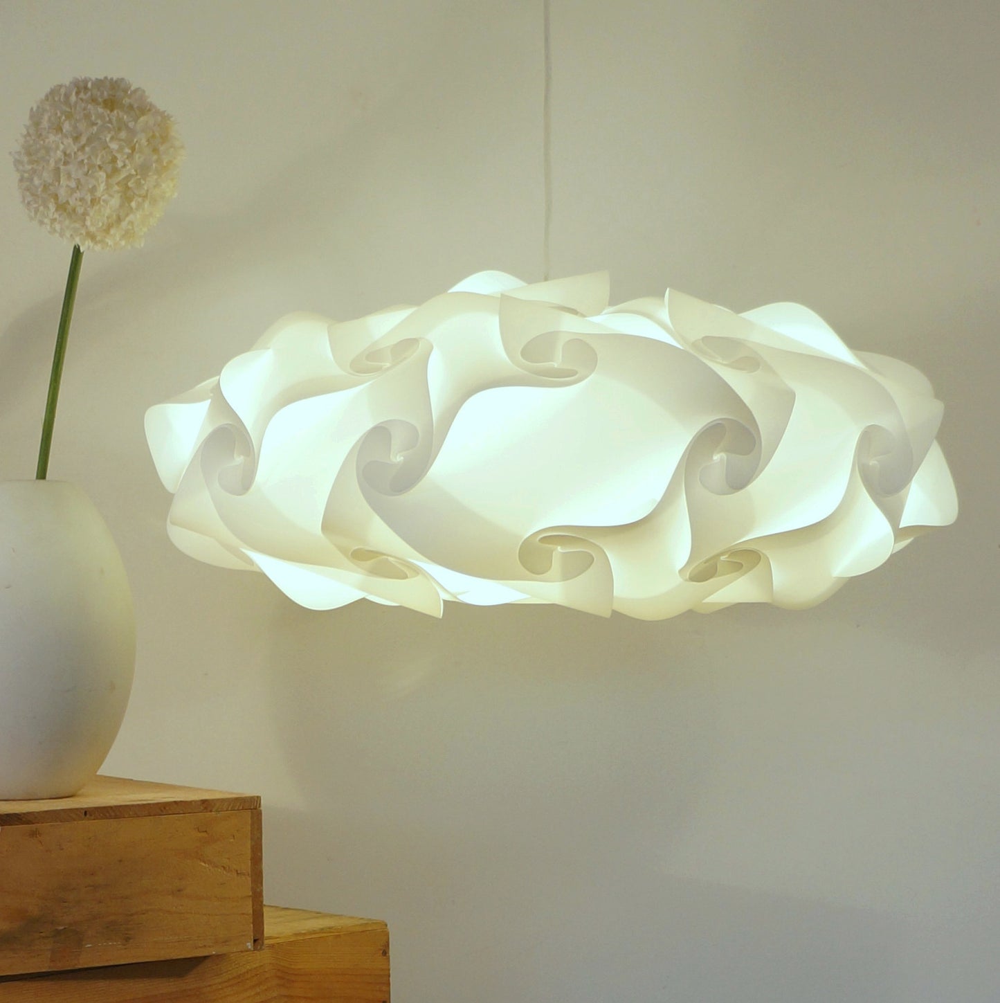 Unique white sculptural lamp shade with flowing organic shapes, placed on a wood surface with a vase and flower in the foreground.