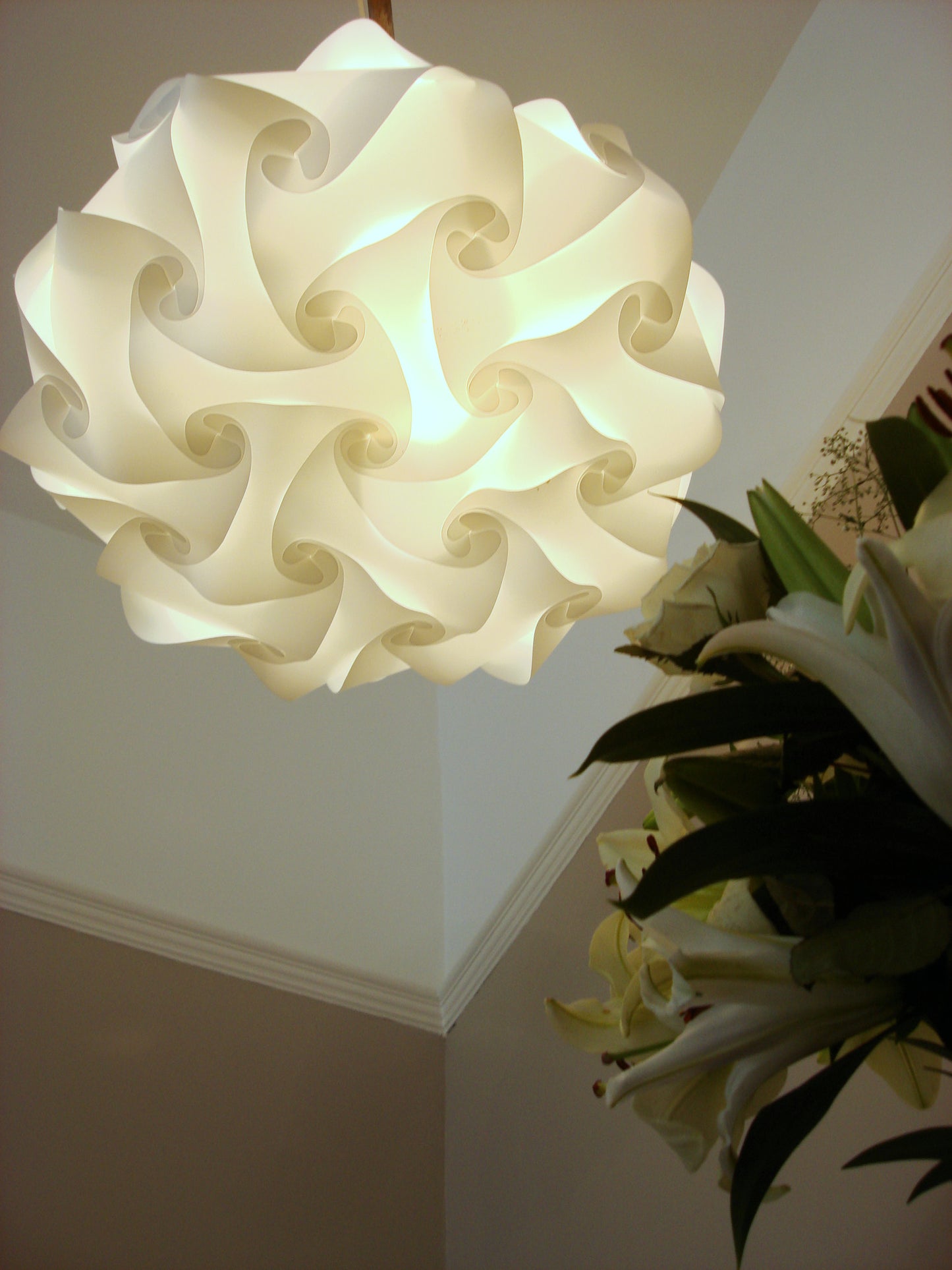 Elegant white swirling lamp shade with intricate design, illuminating a cozy home interior with potted plants in the background.