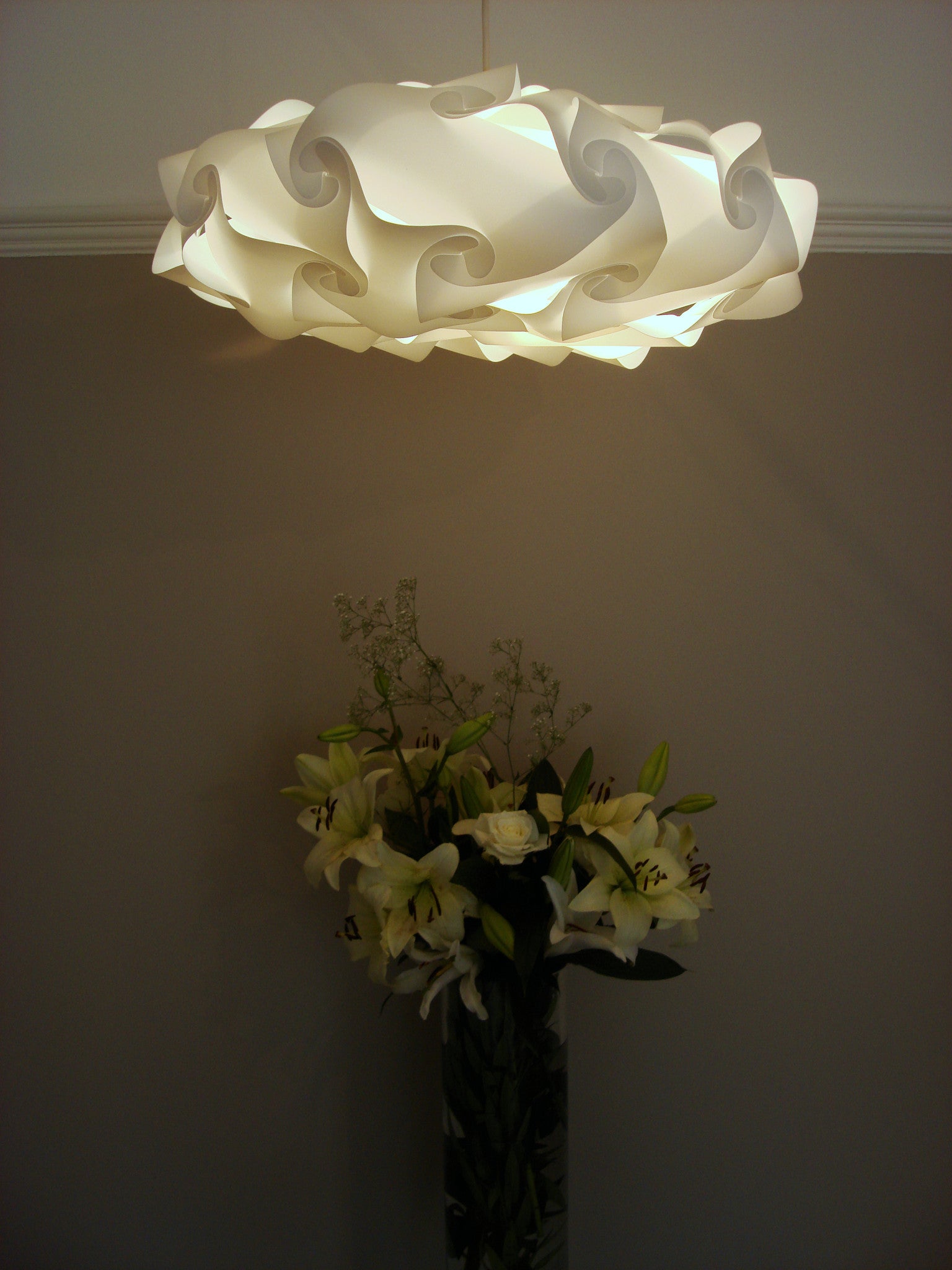 Elegant white lamp shade with intricate swirling design hanging above a vase of fresh white flowers.