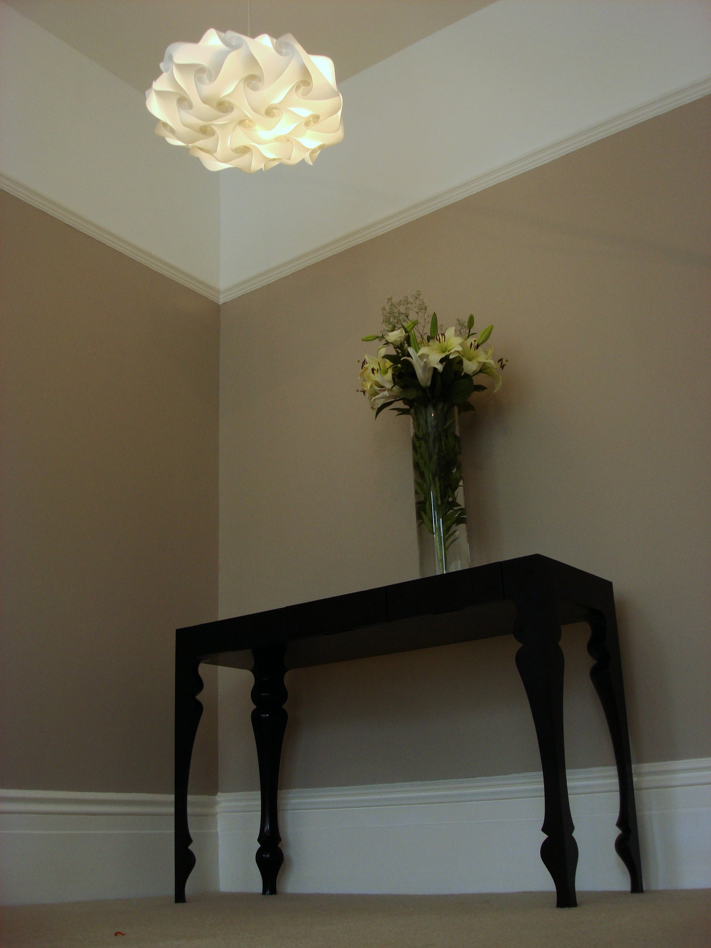Ornate white pendant light fixture above a dark wooden console table with a vase of green and white flowers in a modern home interior.