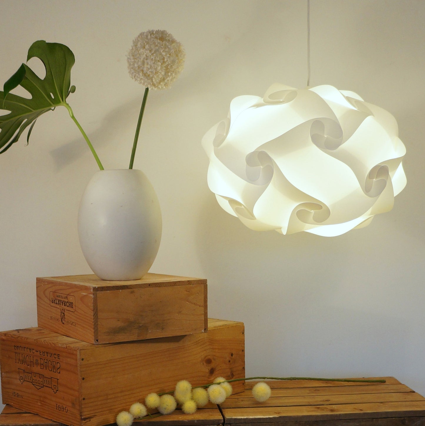 Elegant paper lamp shade suspended from ceiling, set against wooden crates and nature-inspired decor elements like a large monstera leaf and dandelion flower.